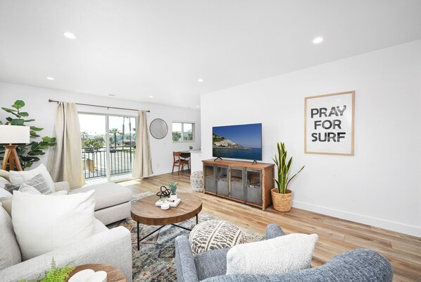 A modern living room with a sofa, coffee table, and a TV displaying a coastal scene. There's a "PRAY FOR SURF" sign on the wall.