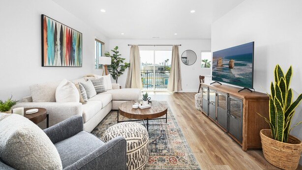A modern living room with a sofa, coffee table, and TV. There's a balcony with a view, a plant, and a dining area with cabinets.
