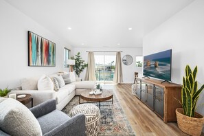 Una sala de estar moderna con sofá, mesa de centro y televisor. Tiene un balcón con vistas y una planta grande.