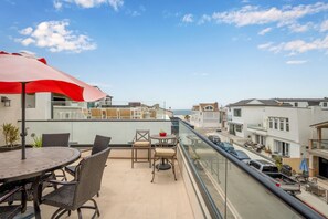 Ein Balkon mit Tisch und Stühlen, einem roten Sonnenschirm und Blick auf das Meer und angrenzende Häuser.