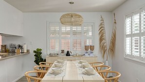 A dining area with a long table set for six, surrounded by wooden chairs, a hanging light fixture, and large windows with white shutters.