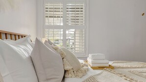 A neatly made bed with white linens and a macrame throw, a window with blinds, and a stack of folded towels on a wooden side table.