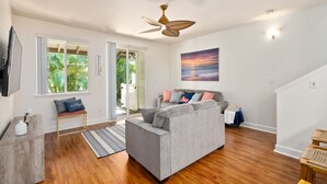 A spacious living room with a grey sofa, wooden flooring, a ceiling fan, and a large window with a view of greenery.