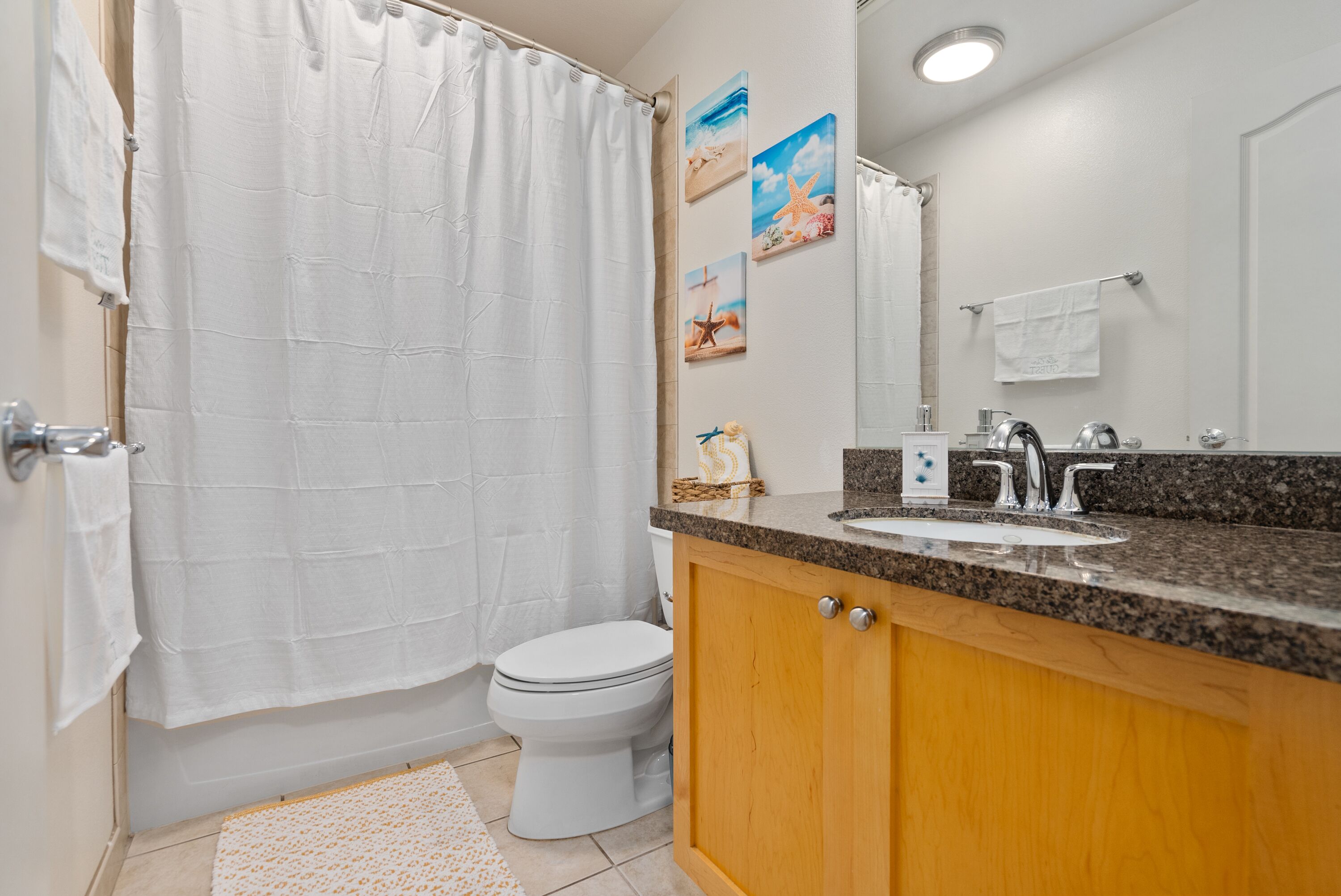 A bathroom with a white toilet, a shower curtain, a sink with a granite countertop, wooden cabinets, and a mirror.
