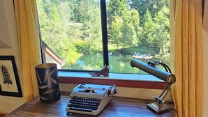 A wooden desk with a typewriter and lamp, a chair, and a view of a forest and lake through a large window.