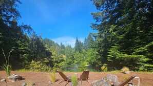 A serene outdoor seating area with two wooden chairs and a large rock centerpiece, surrounded by a forest and a lake.