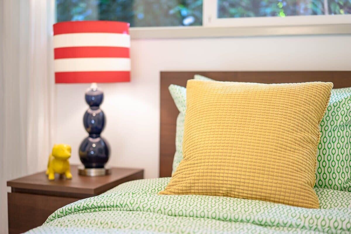 A bed with a green patterned bedspread, a yellow textured pillow, and a red and white striped lamp on a wooden nightstand with a small yellow figurine.