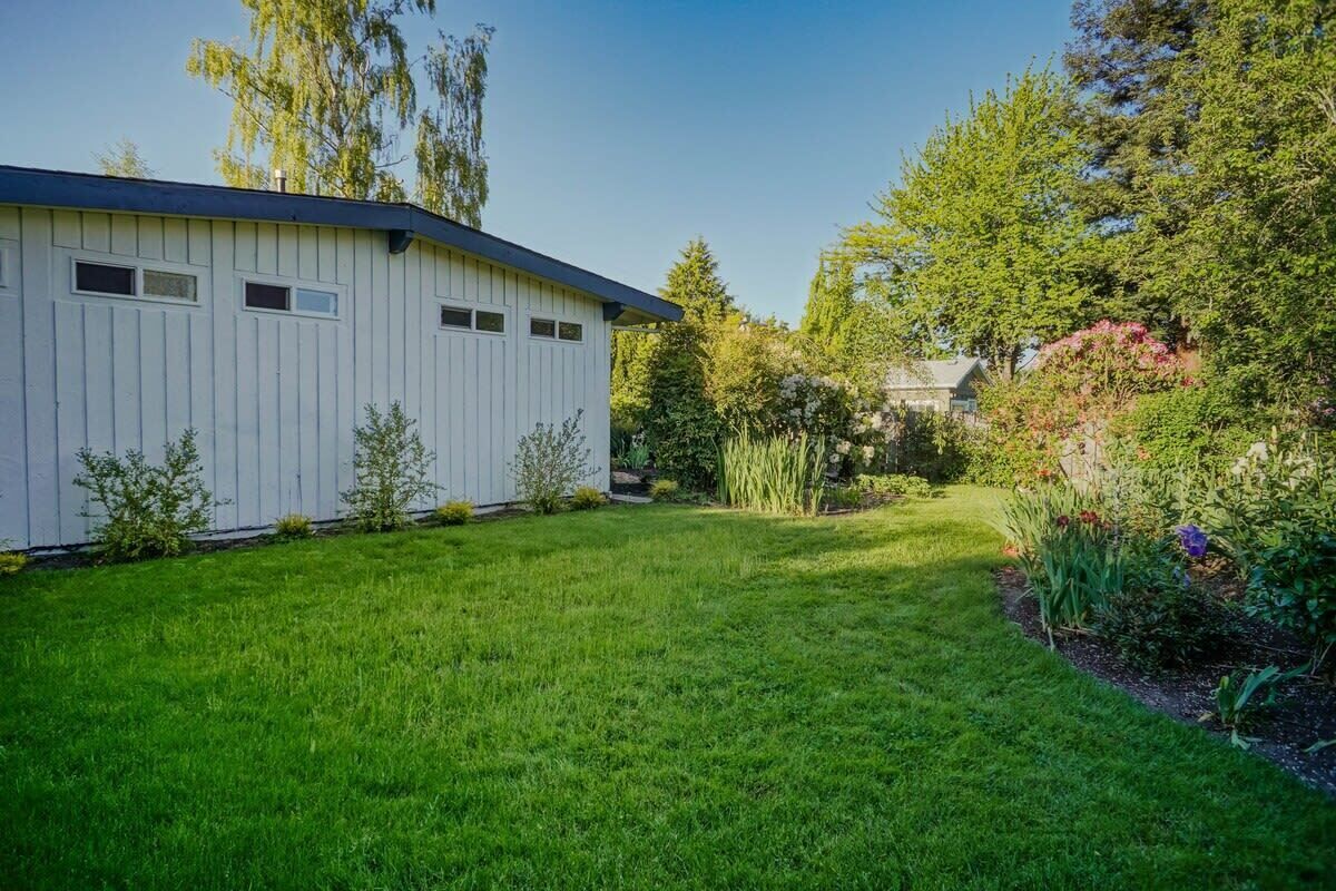 A white building with a sloped roof, surrounded by a well-maintained lawn and various shrubs.