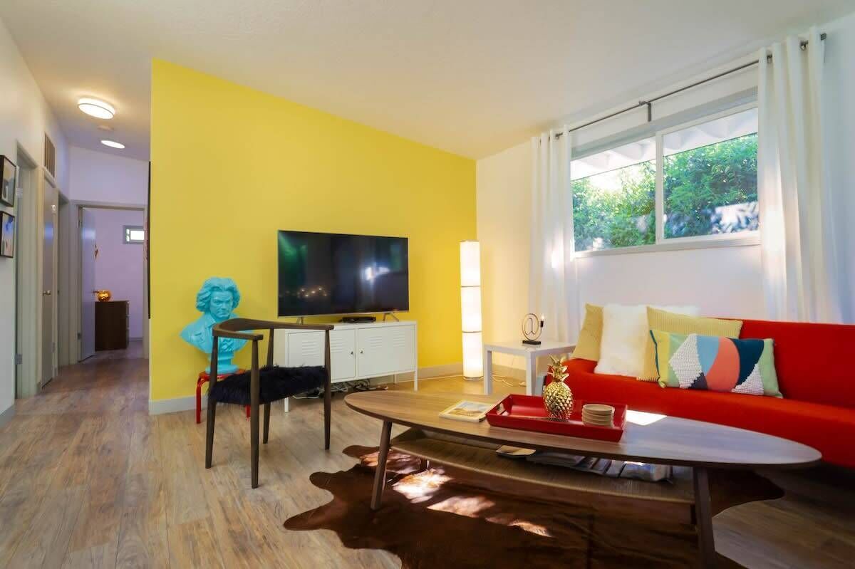 Ein Wohnzimmer mit gelber Wand, einem roten Sofa, einem Fernseher und einem Fenster mit Vorhängen.