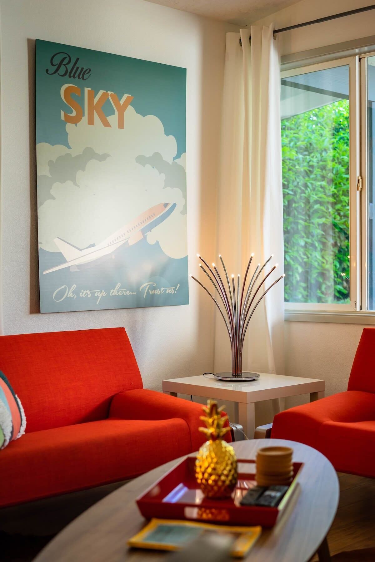 Un salon moderne avec un canapé rouge, une table basse sur laquelle se trouvent un ananas et un plateau, une œuvre d’art accrochée au mur représentant un avion, et une fenêtre donnant sur un paysage verdoyant.