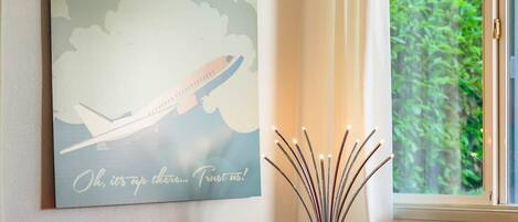 Un salon moderne avec un canapé rouge, une table basse sur laquelle se trouvent un ananas et un plateau, une œuvre d’art accrochée au mur représentant un avion, et une fenêtre donnant sur un paysage verdoyant.