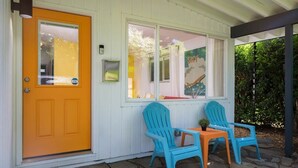 Une maison avec une porte d’un orange vif, des murs extérieurs blancs et une véranda avec deux fauteuils Adirondack bleus et une petite table.