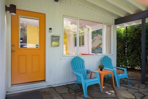 Una casa con una puerta de color naranja brillante, paredes exteriores blancas y un porche con dos sillas Adirondack azules y una mesa pequeña.