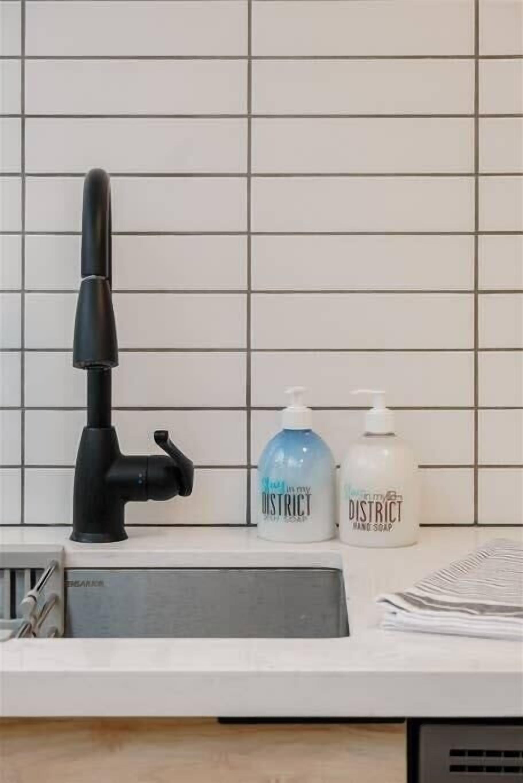 A bathroom sink with a black faucet, two soap dispensers labeled "In my District Hand Soap," and a white tiled wall.