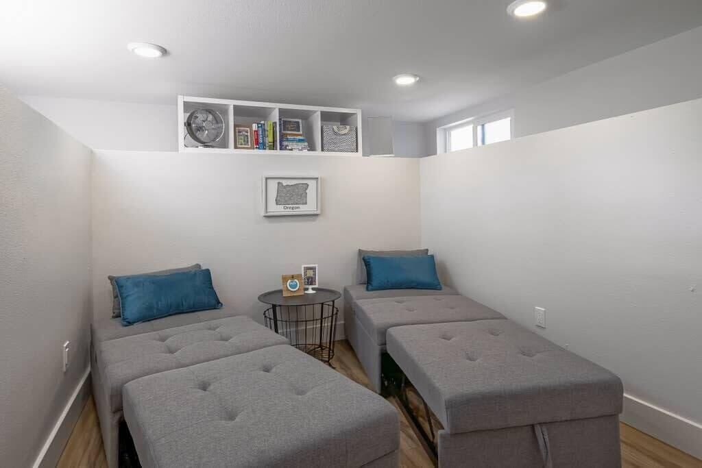Ein kleines Zimmer mit zwei grauen Betten, einem runden Tisch und einer Ablage mit Büchern und einer gerahmten Karte.
