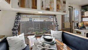 A kitchen with wooden cabinets, a dining table with plates, a window with patterned curtains, and a blue bench. - #StayinMyDistrict Twin Falls Backyard Glamper (Twin Falls)