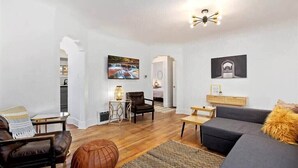 Un salon moderne avec un canapé, des fauteuils, une table basse et une cheminée. Des tableaux encadrés ornent les murs, et une lampe est posée sur une table d’appoint.