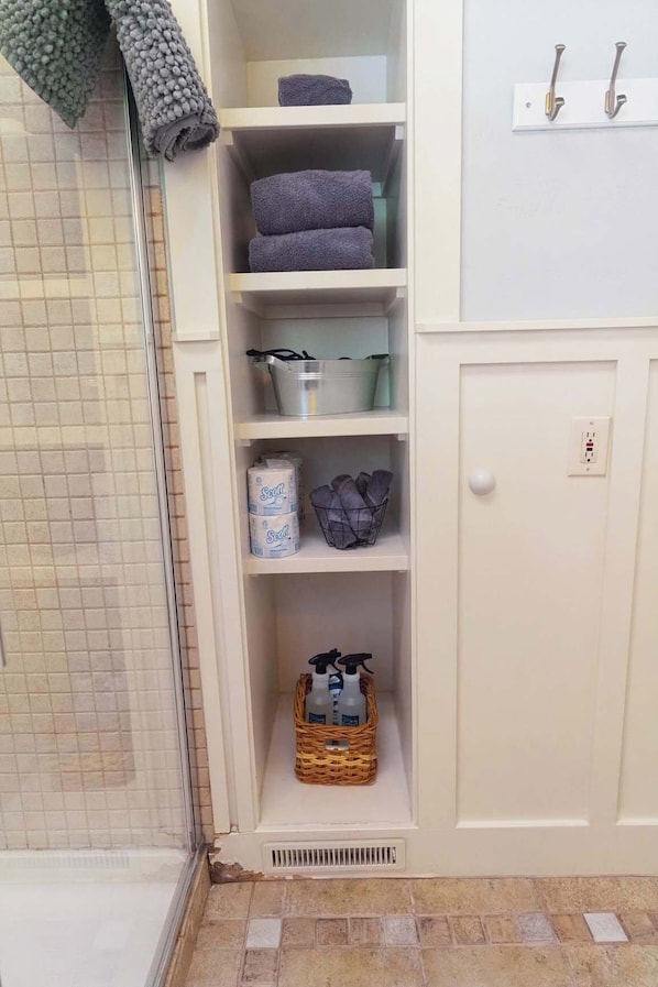 Un baño con suelo de baldosas, zona de ducha y un armario con estantes que contienen toallas, una cesta y productos de limpieza.