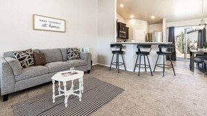 Un salón moderno con un sofá gris, una mesita blanca pequeña y un tapete con estampado blanco y negro. Se observan tres taburetes negros y una zona de cocina al fondo.
