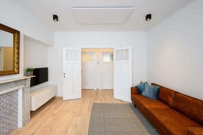 Uma sala de estar moderna com piso de madeira, um sofá marrom, uma TV de tela plana, uma lareira e portas brancas que dão para outro cômodo.