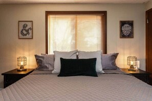 A bedroom with a large bed, two bedside tables with lamps, a window with blinds, and two framed pictures on the walls.