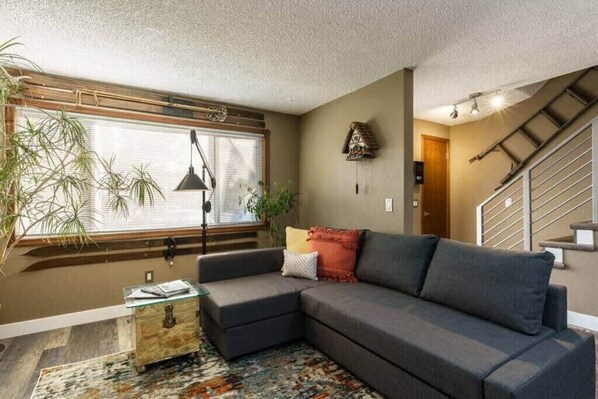 A living room with a sectional sofa, a glass coffee table, a patterned rug, a window with blinds, a wall-mounted birdhouse, and a staircase.