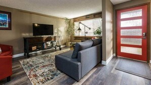A living room with a gray sofa, a red chair, a TV on a wooden cabinet, a red door, and a large window with blinds. - #StayInMyDistrict Eclectic Boise Townhouse near St. Als (Boise)