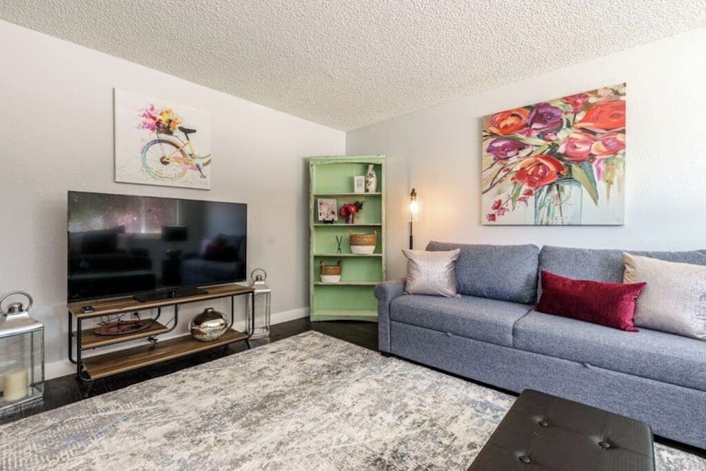Uma sala de estar com uma televisão de tela plana, um aparador verde, um sofá cinza e um quadro com flores.