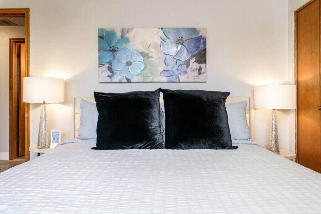 A hotel room with a large bed, two black pillows, white bedding, and a floral painting on the wall.