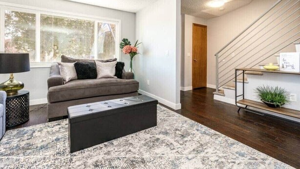 A modern living room with a grey sofa, a black coffee table, and a patterned rug. A staircase with a metal railing leads to a landing with a shelf. - #StayInMyDistrict Cozy Central Boise Townhouse (Boise)