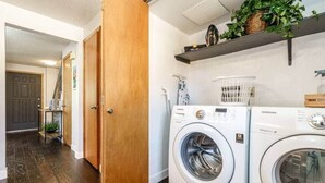 A laundry room with two white front-loading washing machines, a wooden door, a basket on the wall, and a plant on a shelf. - #StayInMyDistrict Cozy Central Boise Townhouse (Boise)