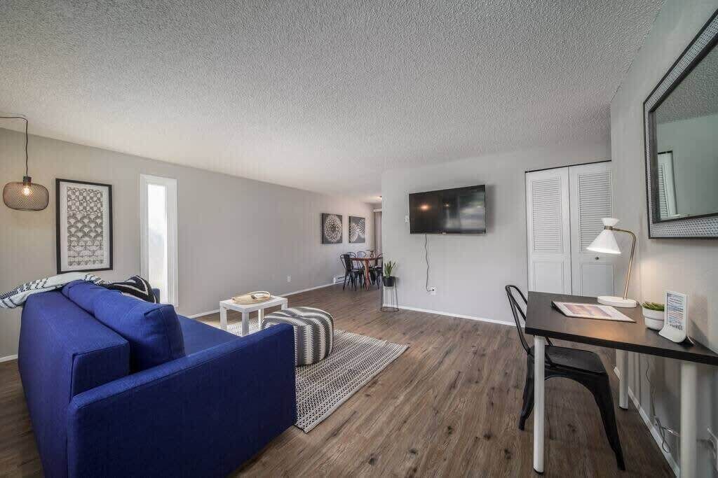 A modern living room with a blue sofa, a dining table with chairs, a flat-screen TV mounted on the wall, and a small side table with a lamp.