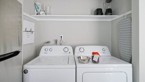 Une buanderie équipée de deux machines à laver à chargement frontal, d’une étagère au-dessus où se trouvent une machine à café et d’autres objets, ainsi que d’un porte-serviettes fixé au mur.