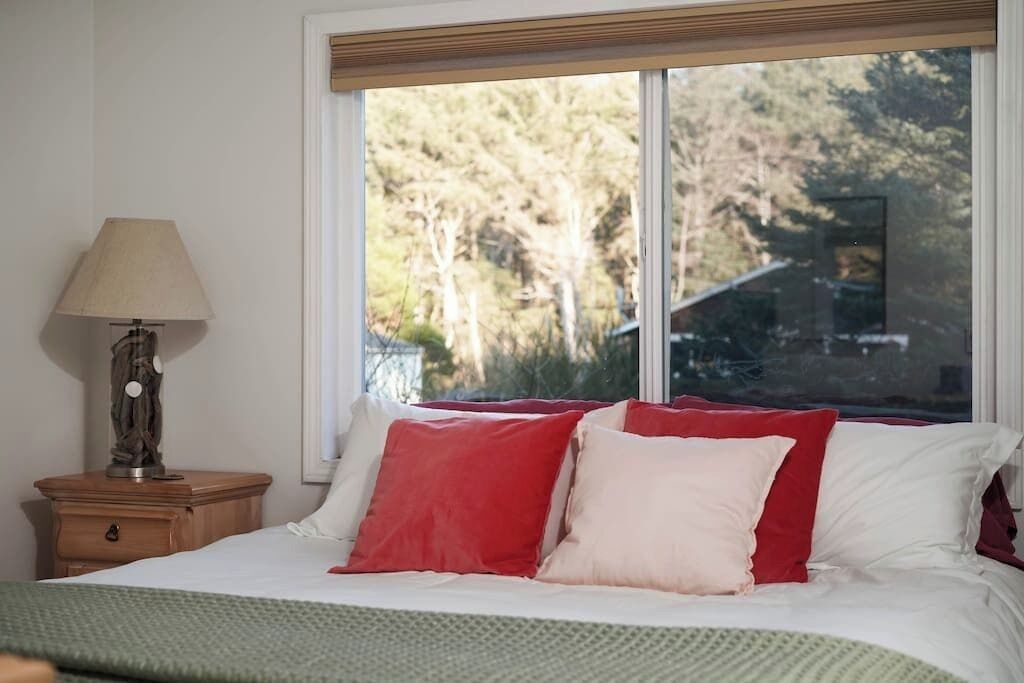 A bedroom with a bed, a nightstand with a lamp, and a window with a view of trees.
