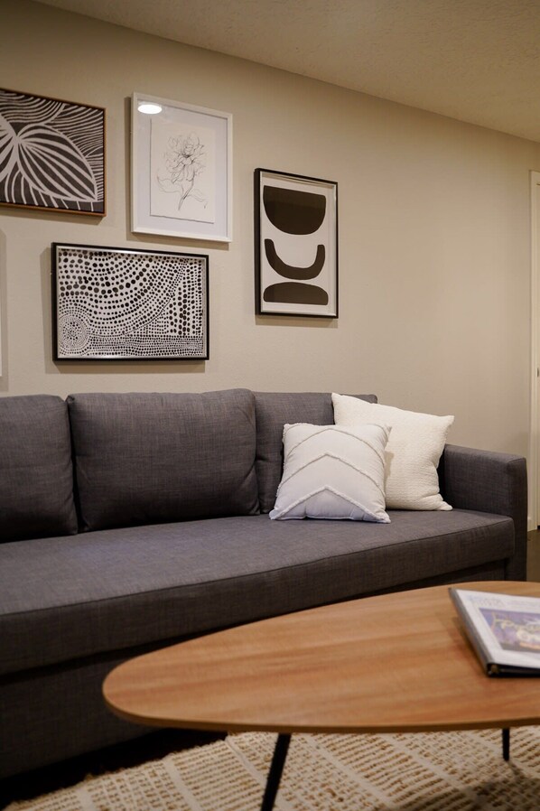 A living room with a gray sofa, two white pillows, and a wooden coffee table. There are three framed artworks on the wall.