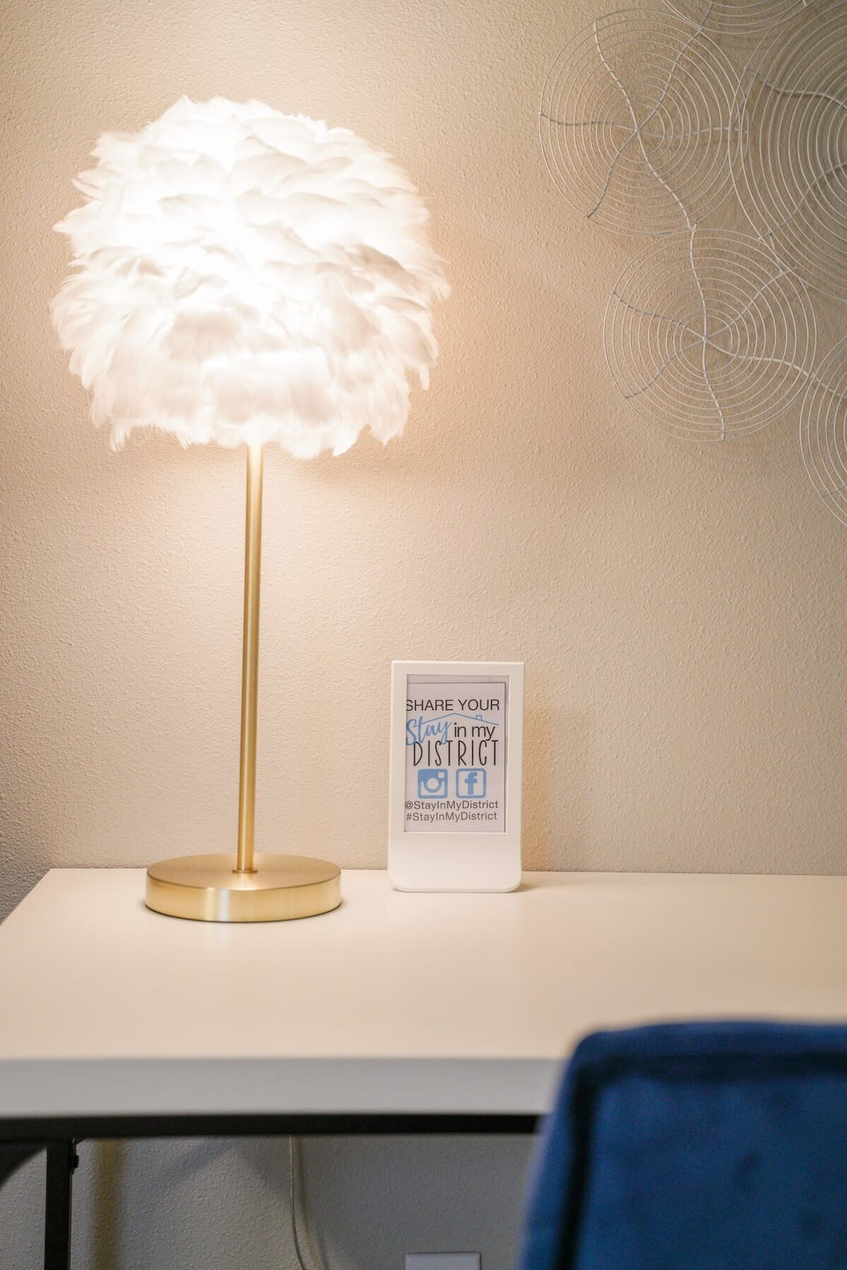 Eine Schreibtischlampe mit einem weißen, federartigen Schirm, ein gerahmtes Schild mit Social-Media-Symbolen und eine Wand mit einem kreisförmigen Muster.