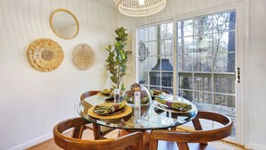 Un coin repas avec une table en verre, des chaises en bois et une vue sur les arbres à travers une grande fenêtre.