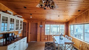 Interior de una cabaña con paredes de madera, una cocina con armarios blancos, una mesa de comedor y ventanas amplias.