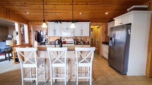 Un interior de madera con una zona de cocina que cuenta con armarios blancos, un refrigerador de acero inoxidable y un conjunto de taburetes de bar.