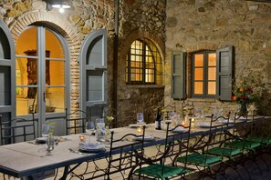 A long dining table set with plates, glasses, and candles, surrounded by green cushioned chairs in a stone-walled room with arched windows and wooden shutters.