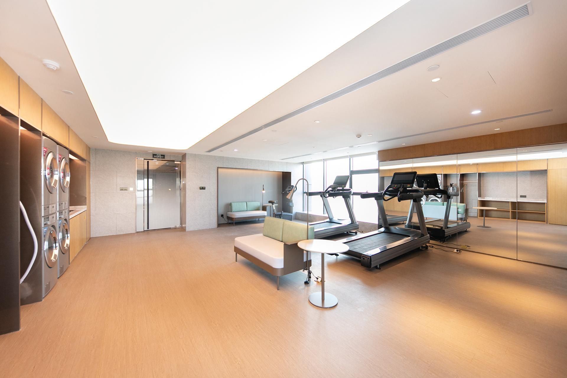 A modern hotel gym with exercise equipment, a seating area, and a large window providing natural light.