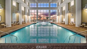 An indoor swimming pool with a view of a sunset through large windows, surrounded by lounge chairs and a brick floor.