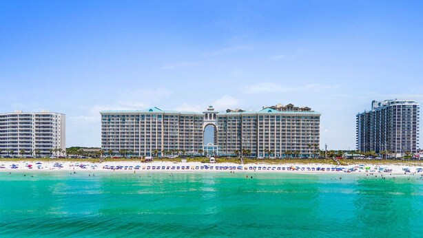 A beachfront hotel with multiple high-rise buildings, a central archway, and a sandy beach with umbrellas.
