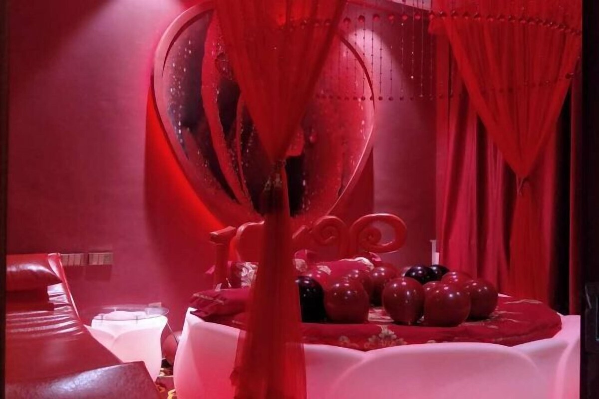 A room with a red theme, including a bed, a table with decorative items, and curtains.