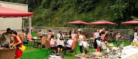 An outdoor event with tables and chairs, a buffet setup, and a grassy area with trees in the background.