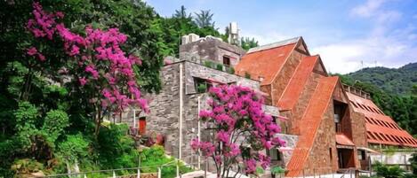 A stone building with a red roof, surrounded by lush greenery and pink flowers.