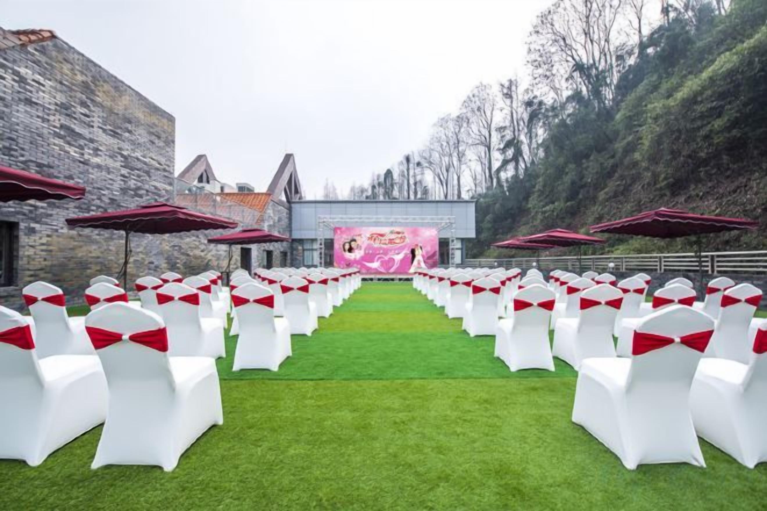 Un salón de bodas al aire libre con hileras de sillas blancas con cintas rojas, un escenario con un telón de fondo rosa y edificios de piedra al fondo.