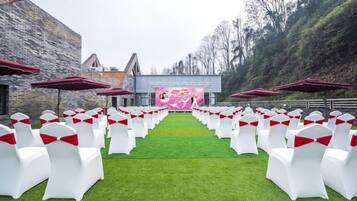 Un salón de bodas al aire libre con hileras de sillas blancas con cintas rojas, un escenario con un telón de fondo rosa y edificios de piedra al fondo.
