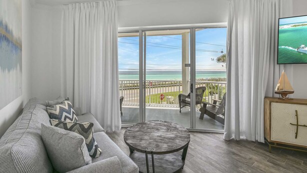 A living room with a sofa, a round coffee table, and a TV on the wall. There is a balcony with a view of the ocean and a stop sign.