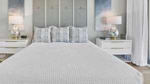 A neatly made bed with a white duvet and patterned pillows, flanked by two bedside tables with lamps and clocks, against a backdrop of a large, neutral-toned headboard and wall art.